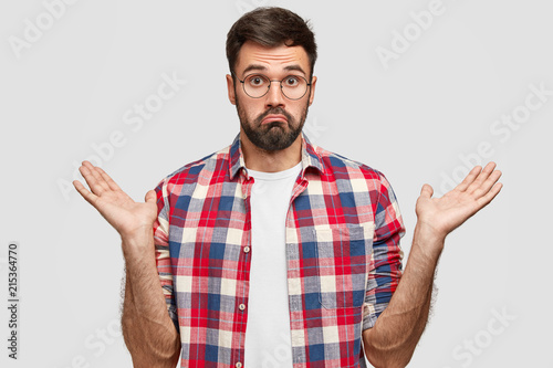 Photo of hesitant unshaven male clasps hands with hesitation, has clueless expression, doubts what to do, dressed in checkered shirt, stands against white background. People and confusement concept