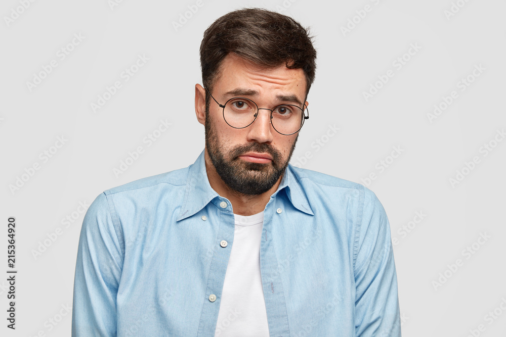 Confused insulted bearded man looks with miserable doubt expression, has to make serious decision in life, wears round eyewear and blue shirt, stands against white wall. People and emotions concept