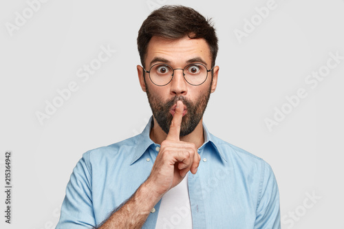 Omg, be silent! Puzzled young male touches lips with index finger, stares at camera, makes hush gesture, afraids someone will tell his secret, has thick bristle, isolated over white background