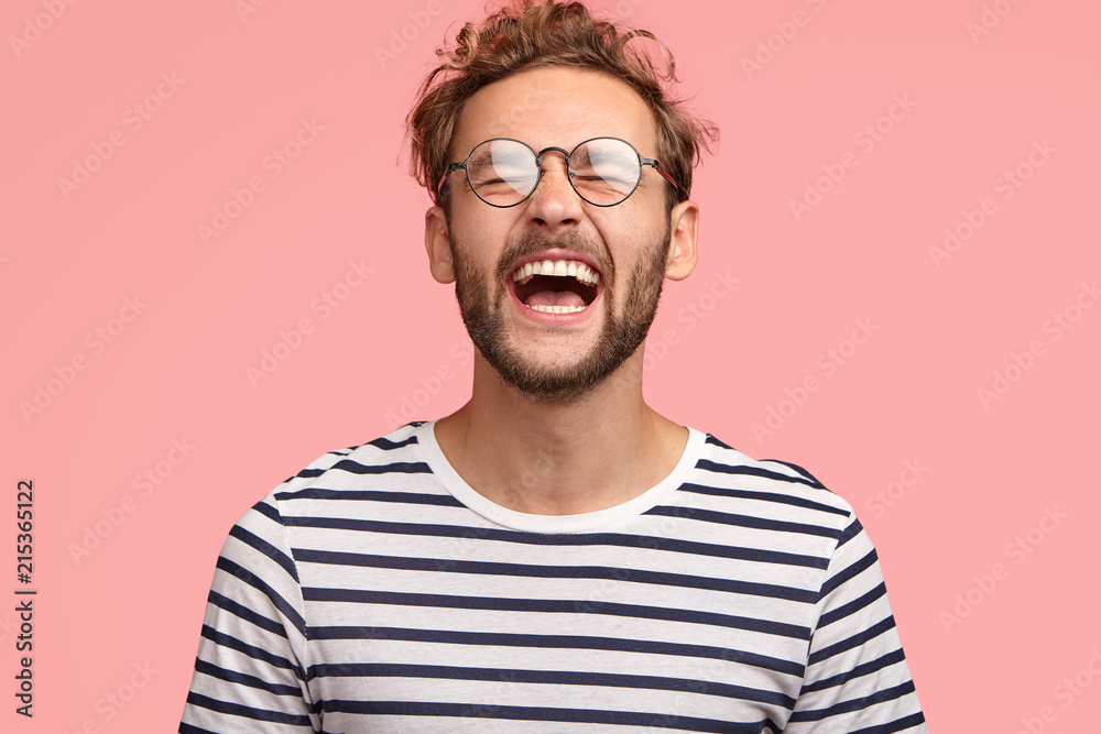 Overjoyed guy laughs joyfully as hears something funny, can`t stop happiness, has carefree lifestyle, wears spectacles, balck and white striped sweater, stands over pink background. Positive emotions Overjoyed guy laughs joyfully as hears something funny, can`t stop happiness, has carefree lifestyle, wears spectacles, balck and white striped sweater, stands over pink background. Positive emotions