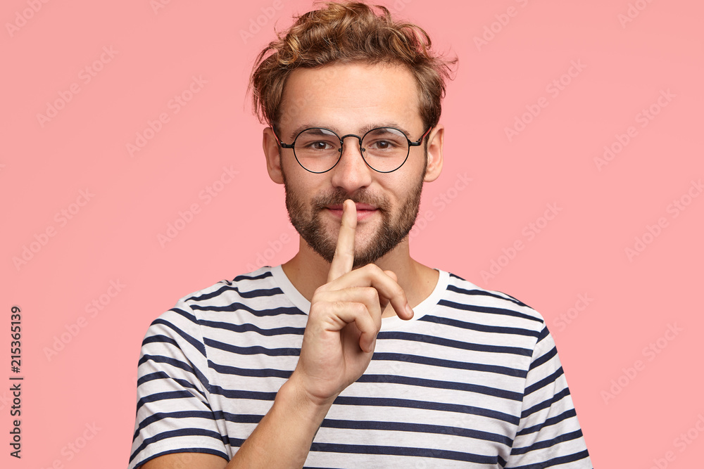 Let`s keep it in secret! Handsome hipster makes shh gesture as spreads rumors, shows hush sign with index finger, dressed casually, isolated over pink background. People, secrecy, conspiracy concept Let`s keep it in secret! Handsome hipster makes shh gesture as spreads rumors, shows hush sign with index finger, dressed casually, isolated over pink background. People, secrecy, conspiracy concept