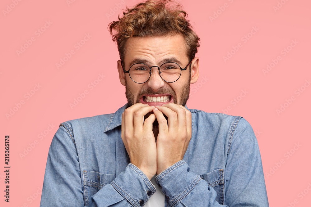 Annoyed young Caucasian male clenches teeth and looks with displeased expression, bites finger nails, looks desperately, feels aversion, wears denim shirt, stands against pink background. Oh no! Annoyed young Caucasian male clenches teeth and looks with displeased expression, bites finger nails, looks desperately, feels aversion, wears denim shirt, stands against pink background. Oh no!