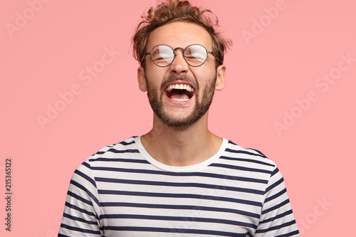 Overjoyed guy laughs joyfully as hears something funny, can`t stop happiness, has carefree lifestyle, wears spectacles, balck and white striped sweater, stands over pink background. Positive emotions