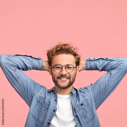 Vertical shot of relaxed carefree man keeps hands behind head, has positive expression, smiles pleasantly, listens something with interest, dressed in fashionable shirt, isolated over pink studio wall photo