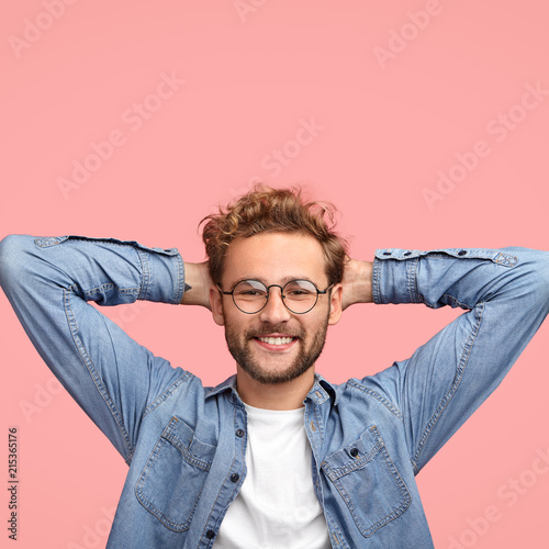Fotografia Vertical shot of relaxed carefree man keeps hands behind head, has positive expr