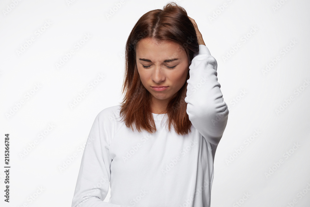 Troubled beautiful young female with dark red hair, keeps hand on back of head, wears white shirt, isolated over white concrete background. Pretty hipster woman has some problems with boss or work. Troubled beautiful young female with dark red hair, keeps hand on back of head, wears white shirt, isolated over white concrete background. Pretty hipster woman has some problems with boss or work.