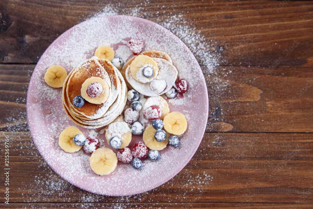 Delicious pancakes with fresh berries sprinkled with powdered sugar in a violet plate on wooden background. A tasty and healthy breakfast of pancakes with raspberries and blueberries. Top view.
