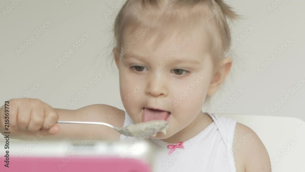 A little girl sitting at a children's table eats porridge with a spoon and looks at the screen of an electronic tablet A little girl sitting at a children's table eats porridge with a spoon and looks at the screen of an electronic tablet