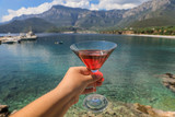 A glass of red martini in the woman hand on the background of a picturesque seascape, summer vacation concept.