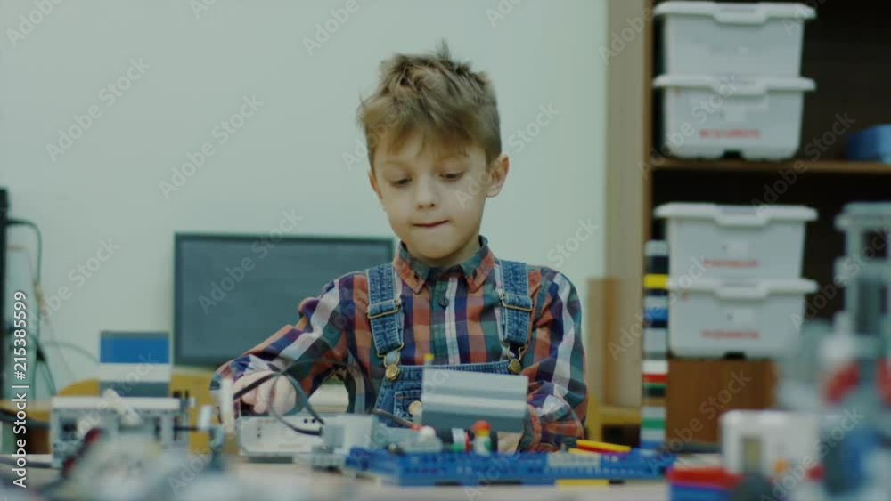 Small funny boy is switching on a robot on a class of robotic technology. He is smiling and waving hands, sitting at table with details Small funny boy is switching on a robot on a class of robotic technology. He is smiling and waving hands, sitting at table with details