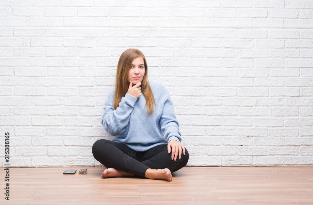 Young adult woman sitting on the floor in autumn over white brick wall looking confident at the camera with smile with crossed arms and hand raised on chin. Thinking positive. Young adult woman sitting on the floor in autumn over white brick wall looking confident at the camera with smile with crossed arms and hand raised on chin. Thinking positive.