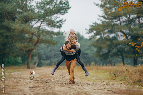 Man carries his wife and daughter on his back at forest path next to dog during walk © Stanislav