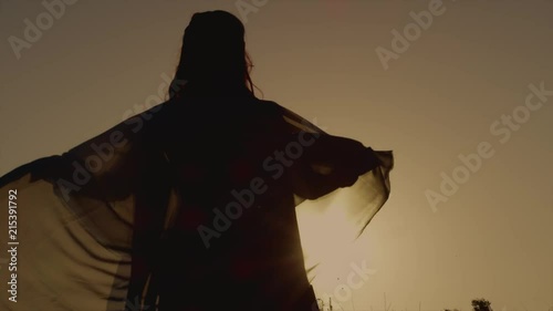 Woman is wrapping in a silk veil and moving over lawn in sunset time. Back view in her dark figure against the sun