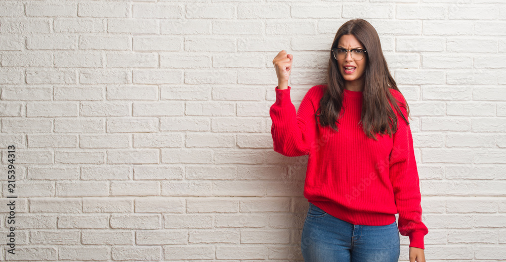 Young brunette woman standing over white brick wall angry and mad raising fist frustrated and furious while shouting with anger. Rage and aggressive concept. Young brunette woman standing over white brick wall angry and mad raising fist frustrated and furious while shouting with anger. Rage and aggressive concept.