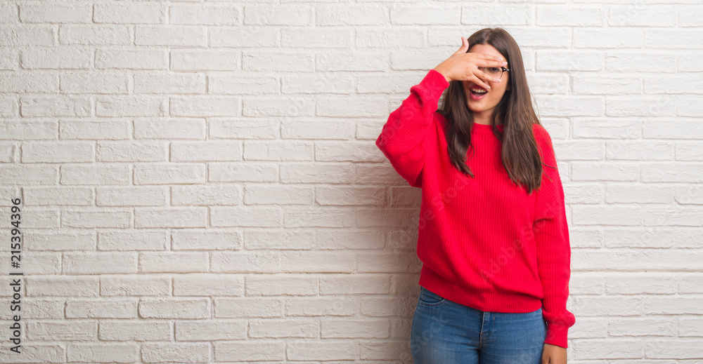 Young brunette woman standing over white brick wall peeking in shock covering face and eyes with hand, looking through fingers with embarrassed expression. Young brunette woman standing over white brick wall peeking in shock covering face and eyes with hand, looking through fingers with embarrassed expression.