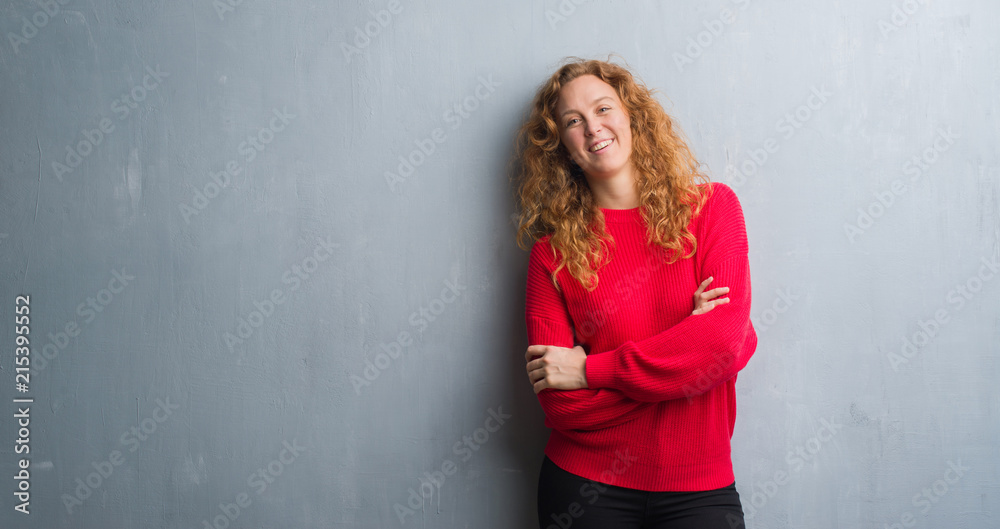 Young redhead woman over grey grunge wall wearing red sweater happy face smiling with crossed arms looking at the camera. Positive person.