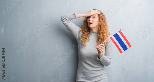 Young redhead woman over grey grunge wall holding flag of Thailand stressed with hand on head, shocked with shame and surprise face, angry and frustrated. Fear and upset for mistake.