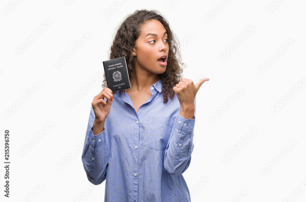 Young hispanic woman holding passport of Italy pointing and showing with thumb up to the side with happy face smiling Young hispanic woman holding passport of Italy pointing and showing with thumb up to the side with happy face smiling