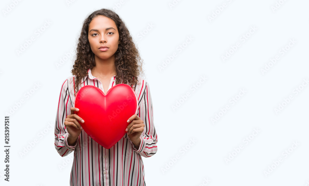 Young hispanic woman in love holding red heart with a confident expression on smart face thinking serious Young hispanic woman in love holding red heart with a confident expression on smart face thinking serious