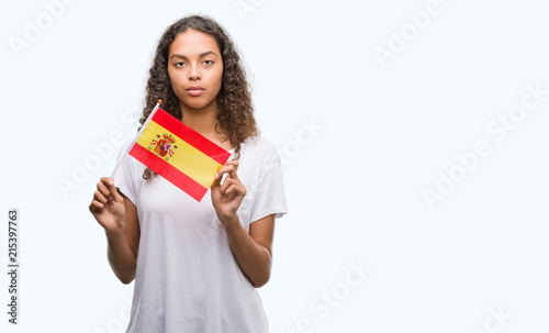 Young hispanic woman holding flag of Spain with a confident expression on smart face thinking serious