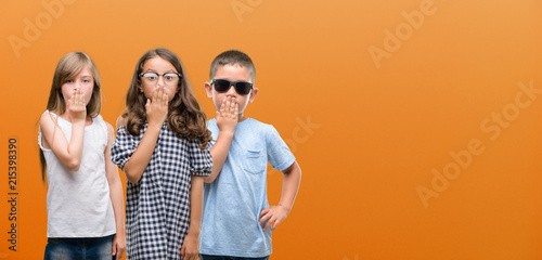 Group of boy and girls kids over orange background cover mouth with hand shocked with shame for mistake, expression of fear, scared in silence, secret concept