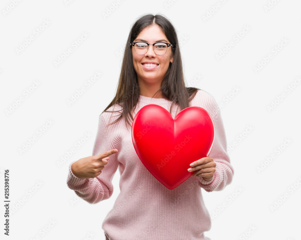 Young hispanic woman holding heart in love very happy pointing with hand and finger