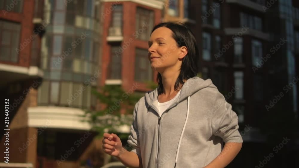 portrait smiling woman running down the street. happy girl enjoy jogging in sunlight. female wearing grey hoodie trains in morning on theckgroung cityscape spring season portrait smiling woman running down the street. happy girl enjoy jogging in sunlight. female wearing grey hoodie trains in morning on theckgroung cityscape spring season