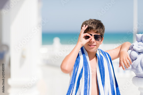 Young child on holidays wearing a navy towel by the beach with happy face smiling doing ok sign with hand on eye looking through fingers