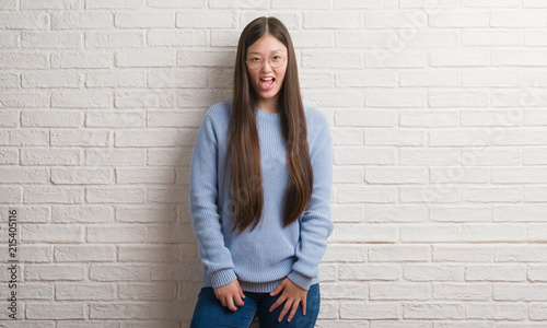 Young Chinise woman over white brick wall sticking tongue out happy with funny expression. Emotion concept.