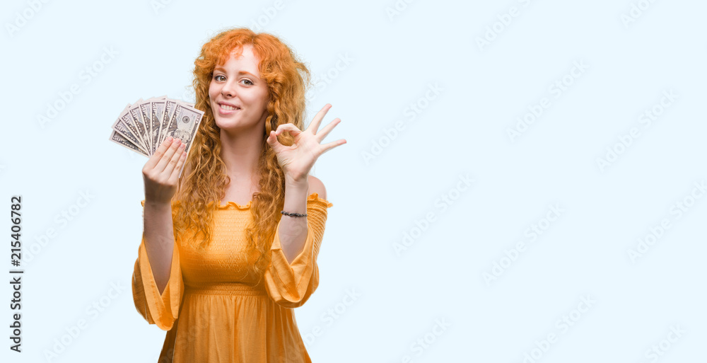 Young redhead woman holding dollars doing ok sign with fingers, excellent symbol Young redhead woman holding dollars doing ok sign with fingers, excellent symbol