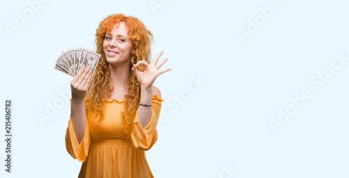 Young redhead woman holding dollars doing ok sign with fingers  excellent symbol
