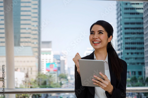 beautiful happy young asian business woman smiling wearing modern black suit cheer up hand with mobile tablet in building city background, network technology, internet, financial, investment concept © Vittaya_25