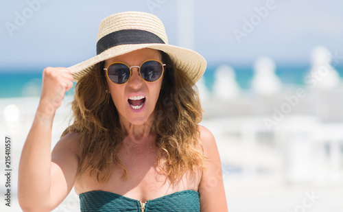 Middle age brunette woman wearing sunglasses by the pool annoyed and frustrated shouting with anger, crazy and yelling with raised hand, anger concept © Krakenimages.com