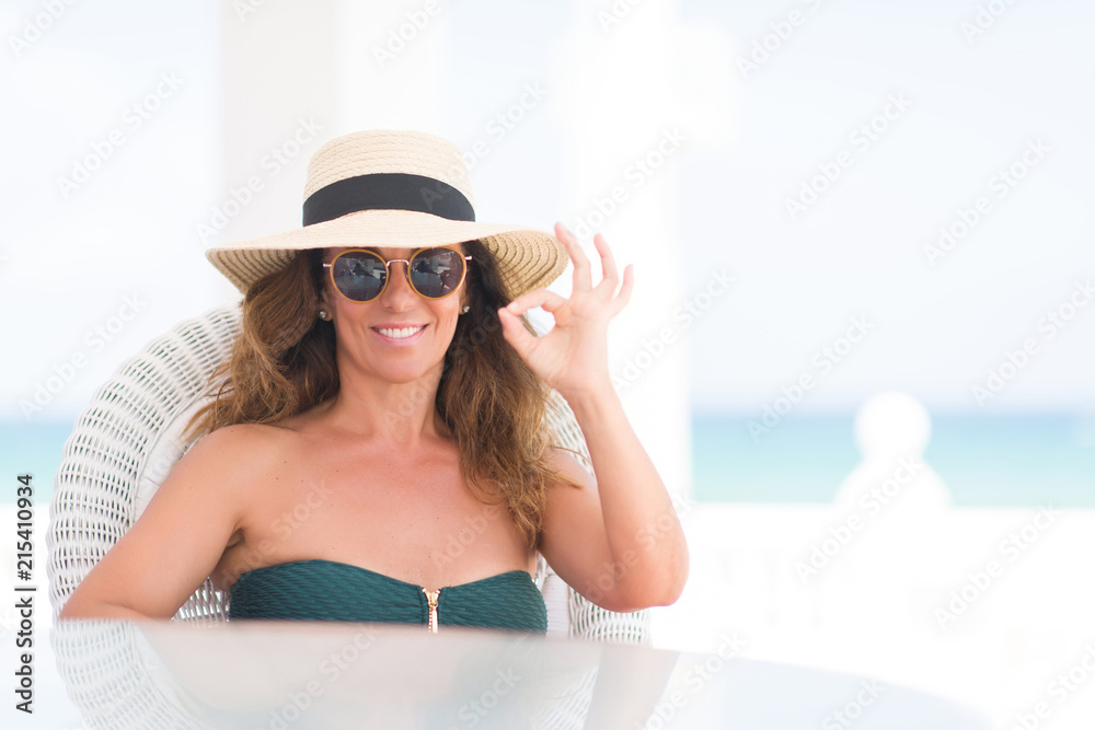 Middle age brunette woman sitting at the table by the beach doing ok sign with fingers, excellent symbol