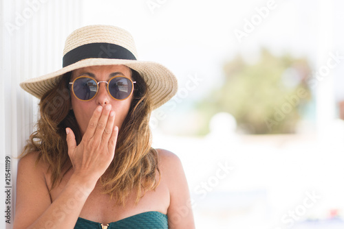 Middle age brunette woman wearing sunglasses and summer hat cover mouth with hand shocked with shame for mistake, expression of fear, scared in silence, secret concept