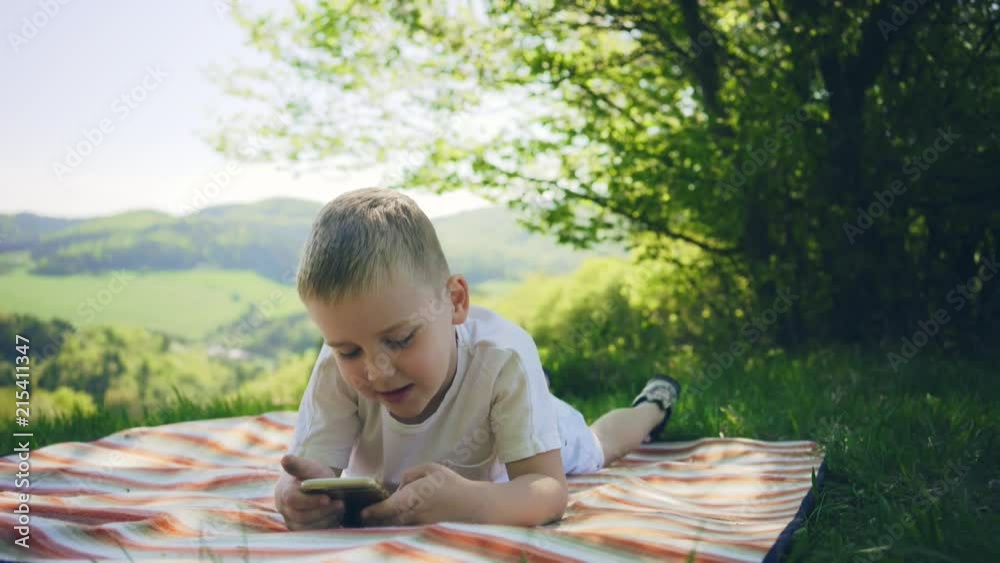 Happy child playing and watching smartphone under sun light. Green field in park at sunny day mountains background Happy child playing and watching smartphone under sun light. Green field in park at sunny day mountains background