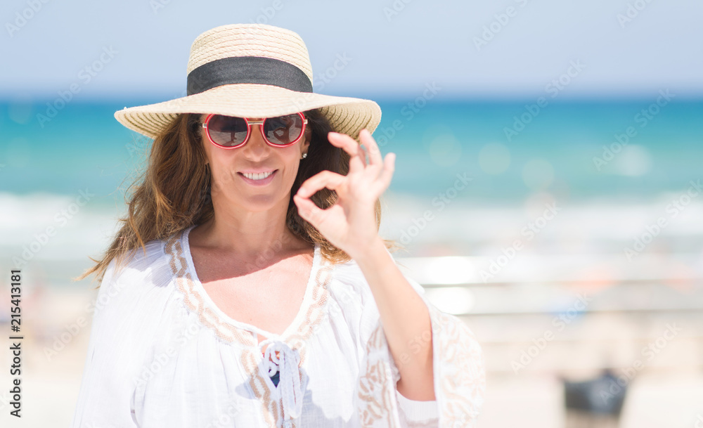 Middle age brunette woman wearing summer hat on vacations by the beach doing ok sign with fingers, excellent symbol Middle age brunette woman wearing summer hat on vacations by the beach doing ok sign with fingers, excellent symbol