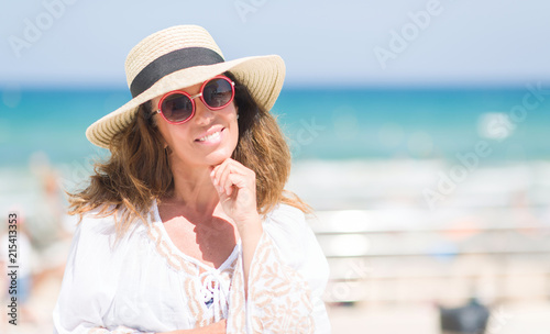 Middle age brunette woman wearing summer hat on vacations by the beach serious face thinking about question, very confused idea