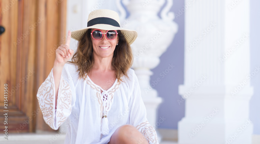 Middle age brunette woman wearing summer hat on vacations surprised with an idea or question pointing finger with happy face, number one