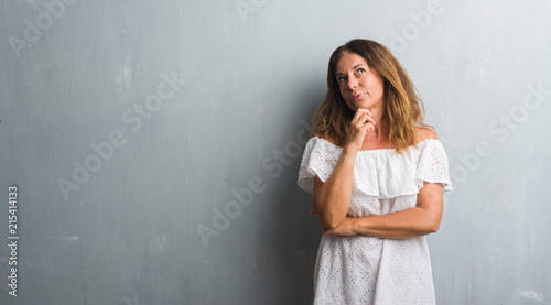 Middle age hispanic woman standing over grey grunge wall with hand on chin thinking about question, pensive expression. Smiling with thoughtful face. Doubt concept. © Krakenimages.com