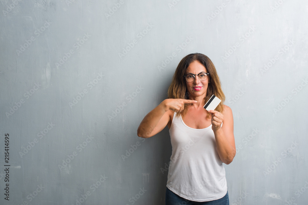Middle age hispanic woman standing over grey grunge wall holding credit card very happy pointing with hand and finger Middle age hispanic woman standing over grey grunge wall holding credit card very happy pointing with hand and finger