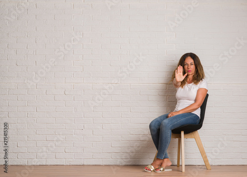 Middle age hispanic woman sitting on chair over white brick walll with open hand doing stop sign with serious and confident expression, defense gesture