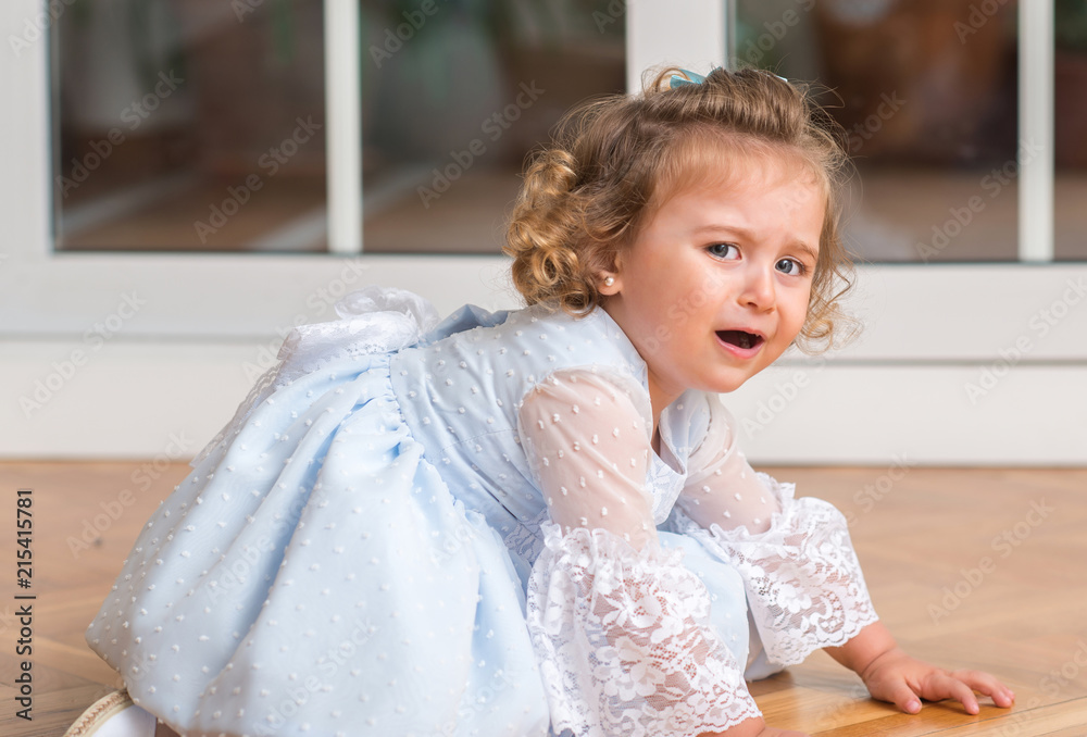 Fototapeta premium Beautiful blonde child in a dress crying with a trantrum sitting on the floor at home