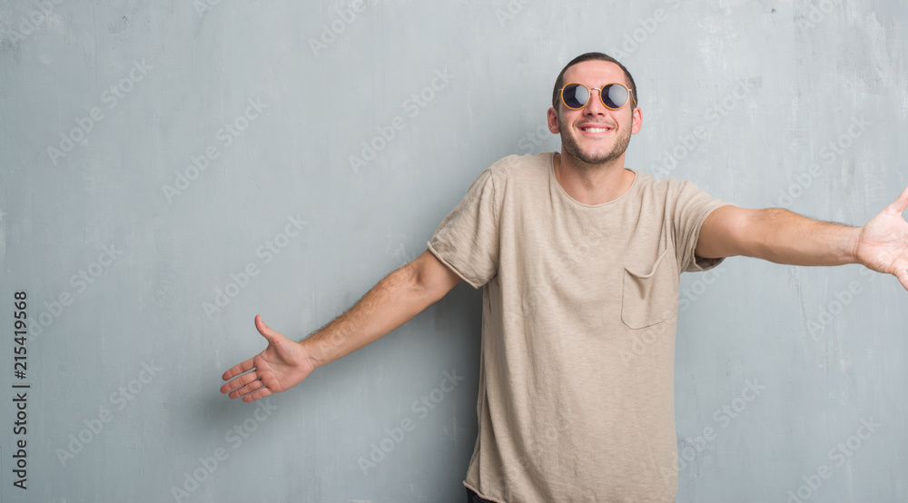 Young caucasian man over grey grunge wall wearing sunglasses looking at the camera smiling with open arms for hug. Cheerful expression embracing happiness. Young caucasian man over grey grunge wall wearing sunglasses looking at the camera smiling with open arms for hug. Cheerful expression embracing happiness.