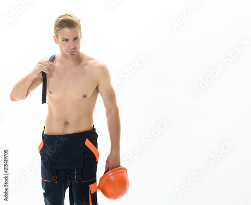 Man in overalls. Worker, repairman, handyman holds hard hat in hand. Strong  builder, industrial worker, mechanic in coveralls. Sexy muscular laborer  with nude torso with protective helmet in overalls. Stock Photo