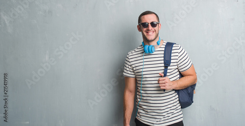 Young caucasian student man over grey grunge wall wearing headphones and backpack with a happy face standing and smiling with a confident smile showing teeth