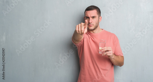 Young caucasian man over grey grunge wall drinking a glass of water pointing with finger to the camera and to you, hand sign, positive and confident gesture from the front © Krakenimages.com