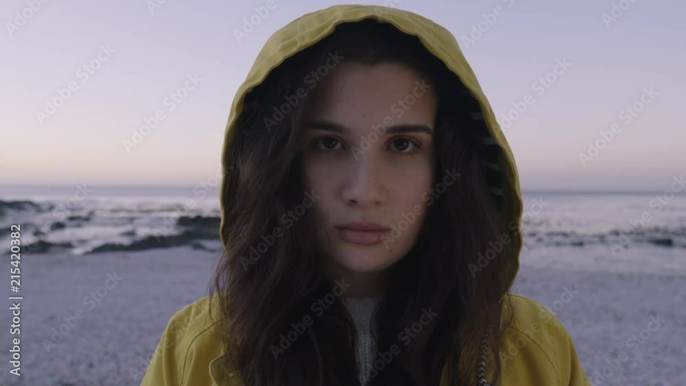 close up portrait of young woman looking serious at camera wearing yellow jacket hood on beautiful calm seaside beach at sunset close up portrait of young woman looking serious at camera wearing yellow jacket hood on beautiful calm seaside beach at sunset