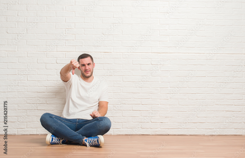Young caucasian man sitting on the floor over white brick wall looking unhappy and angry showing rejection and negative with thumbs down gesture. Bad expression.
