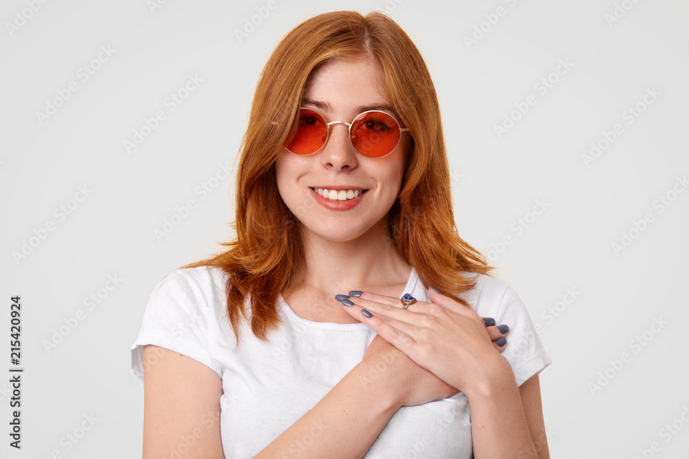 Ginger female with positive smile, keeps hands on chest, expresses good attitude, wears trendy red shades, dressed in casual white t shirt, stands indoor at studio. People and body language concept Ginger female with positive smile, keeps hands on chest, expresses good attitude, wears trendy red shades, dressed in casual white t shirt, stands indoor at studio. People and body language concept